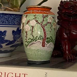 May include: A small, decorative vase with a light green background and a brown tree design. The vase has white flowers and a dark rim. The top rim is orange with gold accents. The vase is sitting on a shelf.