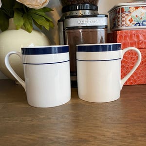 May include: Two white ceramic mugs featuring a dark blue band around the top rim and a thinner dark blue horizontal line below it. Each mug has a curved handle on the side and a smooth, cylindrical body with a glossy finish and visible fine cracks.