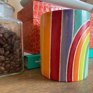 Puede incluir: Una taza cil&iacute;ndrica de cer&aacute;mica con un dise&ntilde;o de arco&iacute;ris. La taza presenta rayas verticales de color naranja, rojo, morado y azul, con un dise&ntilde;o de arco&iacute;ris curvo. La taza est&aacute; sobre una superficie de madera, con un tarro de cristal de granos de caf&eacute; y otros objetos decorativos en el fondo.