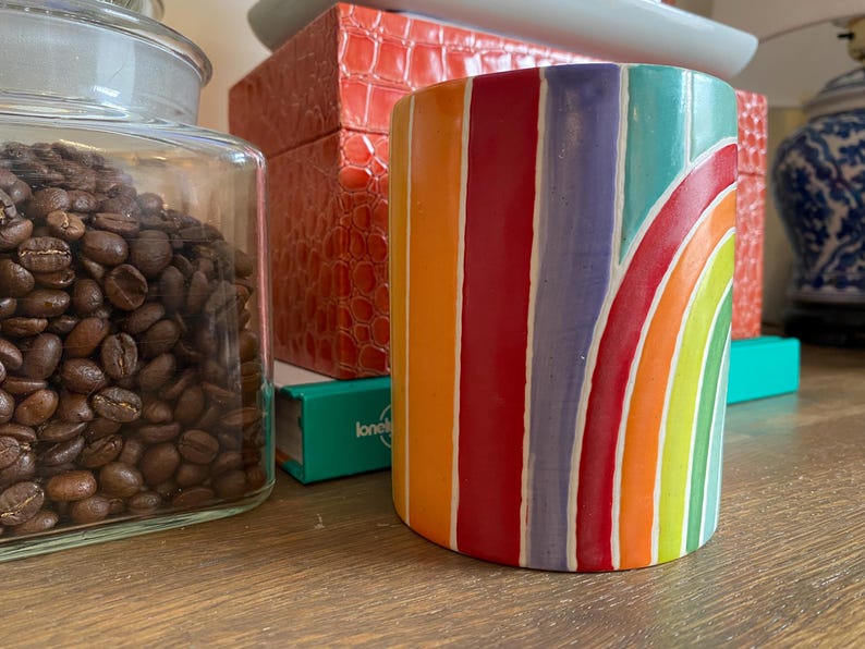 Puede incluir: Una taza cil&iacute;ndrica de cer&aacute;mica con un dise&ntilde;o de arco&iacute;ris. La taza presenta rayas verticales de color naranja, rojo, morado y azul, con un dise&ntilde;o de arco&iacute;ris curvo. La taza est&aacute; sobre una superficie de madera, con un tarro de cristal de granos de caf&eacute; y otros objetos decorativos en el fondo.