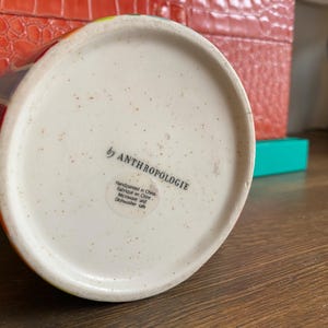 Puede incluir: Una taza de cer&aacute;mica blanca con un dise&ntilde;o de rayas coloridas. La parte inferior de la taza presenta el texto "by ANTHROPOLOGIE" y una peque&ntilde;a etiqueta con texto adicional. La taza est&aacute; pintada a mano y es apta para microondas y lavavajillas.