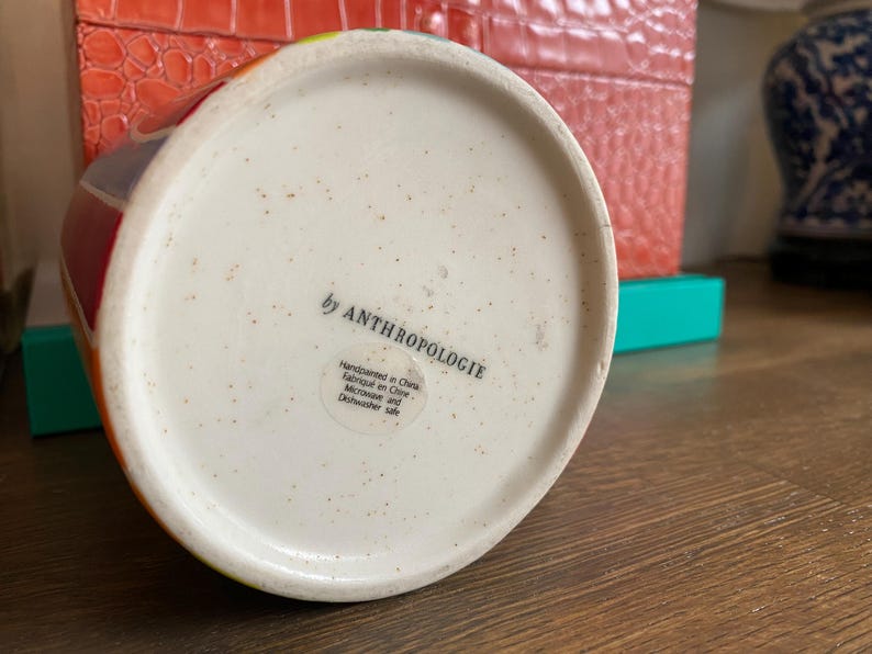 Puede incluir: Una taza de cer&aacute;mica blanca con un dise&ntilde;o de rayas coloridas. La parte inferior de la taza presenta el texto "by ANTHROPOLOGIE" y una peque&ntilde;a etiqueta con texto adicional. La taza est&aacute; pintada a mano y es apta para microondas y lavavajillas.