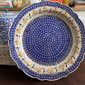 May include: A round ceramic plate with a scalloped edge featuring a dense pattern of small blue and white dots in the center. The rim is decorated with a repeating vine motif of orange flowers and green leaves on a cream-colored background.