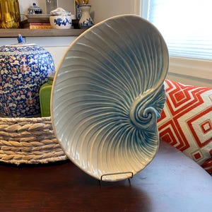 Könnte beinhalten: Eine große, hellblaue dekorative Keramikplatte in Form einer Nautilusmuschel. Die Platte wird auf einem schwarzen Metallständer präsentiert. Das Muscheldesign weist ein Spiralmuster mit einem Farbverlauf von Blau und Weiß auf.