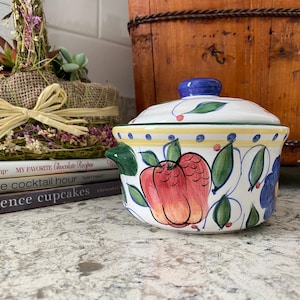 Puede incluir: Una fuente de cerámica blanca con tapa, con manzanas rojas pintadas a mano y hojas verdes. El plato tiene un pomo azul y asas verdes. Una cesta decorativa y libros están en el fondo.