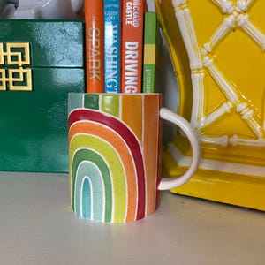 Puede incluir: Una taza de cer&aacute;mica con un dise&ntilde;o de arco&iacute;ris en tonos rojos, naranjas, amarillos, verdes y azules. La taza tiene un asa blanca y est&aacute; llena de libros coloridos. Una caja verde con un dise&ntilde;o geom&eacute;trico dorado y un objeto decorativo amarillo est&aacute;n en el fondo.