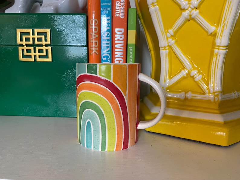Puede incluir: Una taza de cer&aacute;mica con un dise&ntilde;o de arco&iacute;ris en tonos rojos, naranjas, amarillos, verdes y azules. La taza tiene un asa blanca y est&aacute; llena de libros coloridos. Una caja verde con un dise&ntilde;o geom&eacute;trico dorado y un objeto decorativo amarillo est&aacute;n en el fondo.