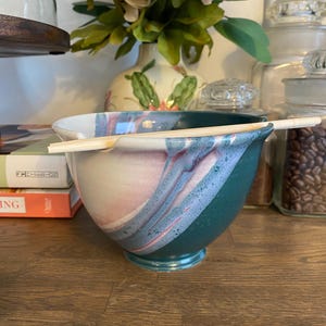 Puede incluir: Un cuenco de cerámica con exterior verde azulado e interior blanco y rosa, con un par de palillos de madera apoyados en el borde. El cuenco tiene una base redonda y una abertura ancha. El cuenco está sobre una superficie de madera.