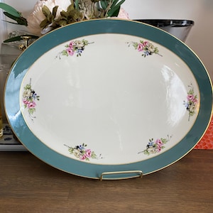 Könnte beinhalten: Eine ovale Servierplatte aus Keramik mit einem breiten blaugrünen Rand und einem dünnen Goldrand. Die Mitte ist cremefarben und mit sechs kleinen Blumensträußen aus rosa Rosen, weißen Blüten und blauen Beeren symmetrisch verziert.