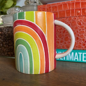 Puede incluir: Una taza de cer&aacute;mica con un dise&ntilde;o de arco&iacute;ris. La taza presenta un asa blanca y un colorido patr&oacute;n de arco&iacute;ris en tonos de rojo, naranja, amarillo, verde y azul. La taza es cil&iacute;ndrica y se encuentra sobre una superficie de madera.
