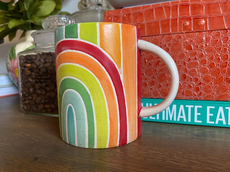 Puede incluir: Una taza de cer&aacute;mica con un dise&ntilde;o de arco&iacute;ris. La taza presenta un asa blanca y un colorido patr&oacute;n de arco&iacute;ris en tonos de rojo, naranja, amarillo, verde y azul. La taza es cil&iacute;ndrica y se encuentra sobre una superficie de madera.