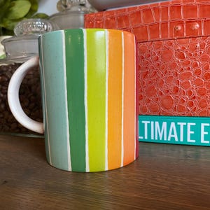 Puede incluir: Una taza de cer&aacute;mica con rayas verticales en tonos verdes, amarillos y naranjas, separadas por l&iacute;neas blancas. La taza tiene un asa blanca. Un tarro de granos de caf&eacute; y un libro con el texto "ULTIMATE EATS" est&aacute;n en el fondo, sobre una superficie de madera.