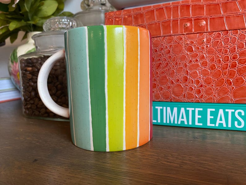 Puede incluir: Una taza de cer&aacute;mica con rayas verticales en tonos verdes, amarillos y naranjas, separadas por l&iacute;neas blancas. La taza tiene un asa blanca. Un tarro de granos de caf&eacute; y un libro con el texto "ULTIMATE EATS" est&aacute;n en el fondo, sobre una superficie de madera.