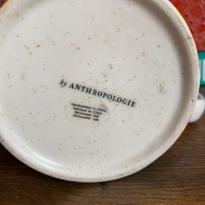 Puede incluir: Vista inferior de una taza de cer&aacute;mica con interior blanco moteado. El texto "by ANTHROPOLOGIE" est&aacute; impreso en negro. Una etiqueta indica que la taza est&aacute; pintada a mano en China, apta para microondas y lavavajillas.