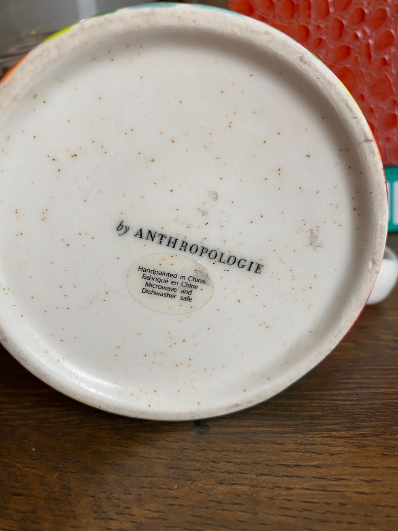Puede incluir: Vista inferior de una taza de cer&aacute;mica con interior blanco moteado. El texto "by ANTHROPOLOGIE" est&aacute; impreso en negro. Una etiqueta indica que la taza est&aacute; pintada a mano en China, apta para microondas y lavavajillas.