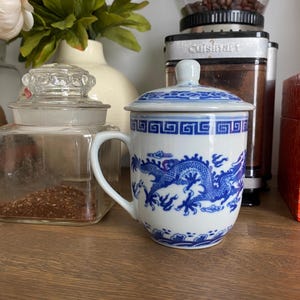 Op de afbeelding: Een witte keramische mok met een blauw drakenpatroon, een bijpassend deksel en een handvat. De mok heeft een traditioneel blauw-wit ontwerp met een gedetailleerd drakenmotief rondom het lichaam en een geometrisch randpatroon bij de opening.