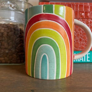 Puede incluir: Una taza de cer&aacute;mica con un dise&ntilde;o de arco&iacute;ris en tonos rojos, naranjas, amarillos, verdes y azules. La taza tiene un asa blanca y est&aacute; sobre una superficie de madera. Un tarro de cristal lleno de granos de caf&eacute; est&aacute; en el fondo.