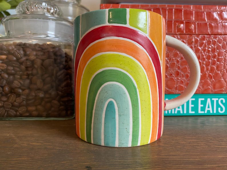 Puede incluir: Una taza de cer&aacute;mica con un dise&ntilde;o de arco&iacute;ris en tonos rojos, naranjas, amarillos, verdes y azules. La taza tiene un asa blanca y est&aacute; sobre una superficie de madera. Un tarro de cristal lleno de granos de caf&eacute; est&aacute; en el fondo.