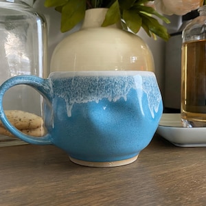 May include: A ceramic mug with a rounded, dimpled body and a curved handle. The mug features a bright blue base color that transitions into a white, drippy glaze effect along the top rim, creating a textured appearance against the smooth surface.