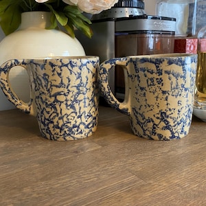 Op de afbeelding: Twee keramische mokken met een crèmekleurige basis en een donkerblauw gespikkeld, met spons aangebracht patroon. Elke mok heeft een gebogen handvat aan de zijkant en een cilindrisch lichaam. De mokken staan naast elkaar en tonen de blauwe afwerking.