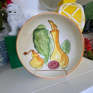 May include: A decorative ceramic serving bowl with a hand-painted design of vegetables in shades of green, yellow, and orange. The bowl has an orange rim and is displayed on a metal stand. The bowl is a unique piece of kitchen decor.
