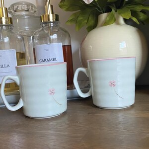 May include: Two off-white ceramic mugs with a subtle horizontal ribbed texture and a small pink flower graphic on the front. Each mug features a matching pink rim and a curved handle, presenting a clean, simple design for daily beverage consumption.