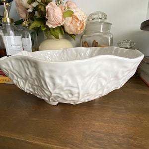 May include: A white ceramic bowl with a textured surface resembling overlapping cabbage leaves. The bowl features a scalloped, wavy rim and is supported by small feet at the base. The exterior has a glossy finish with intricate leaf vein details.