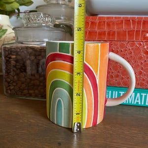 Puede incluir: Una taza de cer&aacute;mica con dise&ntilde;o de arco&iacute;ris, de aproximadamente 10 cm de altura. La taza presenta un asa blanca y un patr&oacute;n colorido de rayas arqueadas en tonos naranja, amarillo, verde y azul. La taza est&aacute; sobre una superficie de madera.