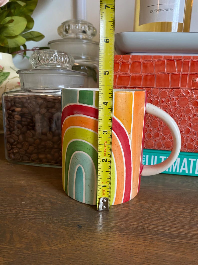Puede incluir: Una taza de cer&aacute;mica con dise&ntilde;o de arco&iacute;ris, de aproximadamente 10 cm de altura. La taza presenta un asa blanca y un patr&oacute;n colorido de rayas arqueadas en tonos naranja, amarillo, verde y azul. La taza est&aacute; sobre una superficie de madera.
