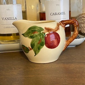 May include: A cream-colored ceramic creamer pitcher featuring a hand-painted design of a single red apple with green leaves on a brown branch. The pitcher has a curved spout and a brown handle shaped like a textured tree branch or twig.