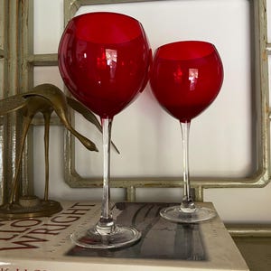 May include: Two wine glasses with deep red, rounded bowls and clear, slender stems. The glasses feature a smooth, polished finish and sit on circular, transparent bases. The design is simple and functional, highlighting the contrast between the vibrant red glass and the clear stem and base components.