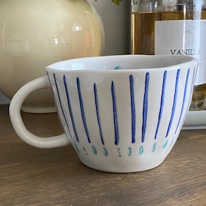 May include: A white ceramic mug with a rounded handle, featuring vertical dark blue painted stripes around the exterior and a row of small light blue dots near the base. The mug has a slightly irregular, hand-formed shape with a smooth finish.