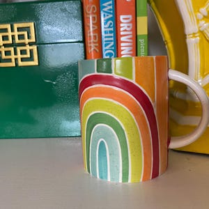 Puede incluir: Una taza de cer&aacute;mica con un dise&ntilde;o de arco&iacute;ris en tonos rojos, naranjas, amarillos, verdes y azules. La taza tiene un asa blanca y est&aacute; colocada frente a una caja verde y un objeto decorativo amarillo.