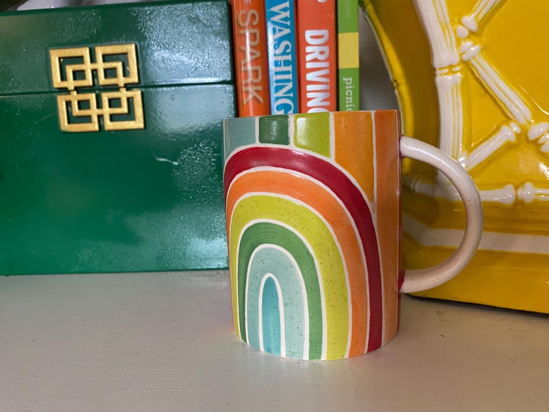 Puede incluir: Una taza de cer&aacute;mica con un dise&ntilde;o de arco&iacute;ris en tonos rojos, naranjas, amarillos, verdes y azules. La taza tiene un asa blanca y est&aacute; colocada frente a una caja verde y un objeto decorativo amarillo.