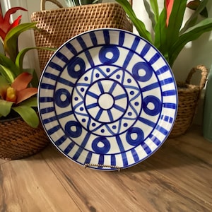 May include: A round ceramic plate featuring a white base with a bold blue hand-painted pattern. The design includes a central sunburst motif surrounded by concentric circles, triangles, and a series of vertical stripes along the outer rim.