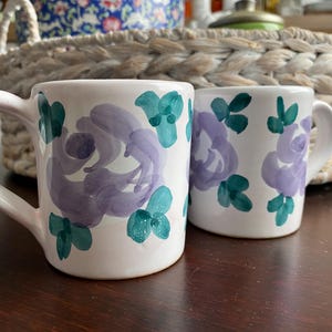 Può includere: Due tazze in ceramica bianca con un motivo floreale dipinto a mano. Ogni tazza presenta grandi fiori color viola tenue, accentuati da piccole foglie verde acqua sparse sulla superficie esterna, completate da un manico ricurvo.