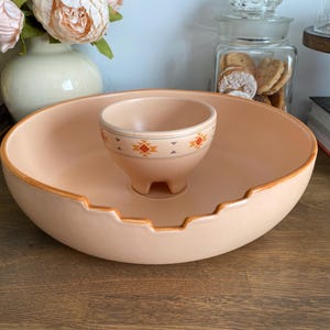 May include: A peach-colored ceramic chip and dip serving set featuring a large, round, shallow bowl with a notched rim and a smaller, integrated central bowl. The smaller bowl is decorated with a geometric pattern in orange, red, and gray.
