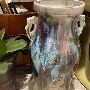 May include: A ceramic vase featuring a wide, flared rim and two side handles. The exterior surface displays a multi-colored glaze with swirling patterns of deep blue, burgundy, white, and green, creating a glossy, marbled appearance throughout.
