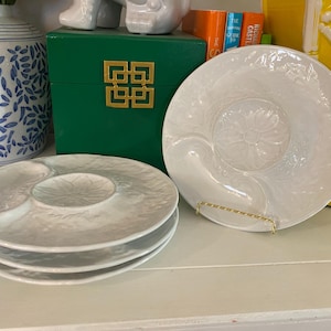 Könnte beinhalten: Ein Stapel von drei weißen, strukturierten Servierschalen mit drei Fächern. Eine vierte Schale mit floralem Design steht auf einem goldenen Ständer. Die Schalen stehen auf einer weißen Oberfläche, mit einer grünen Box und einer blau-weißen Vase im Hintergrund.
