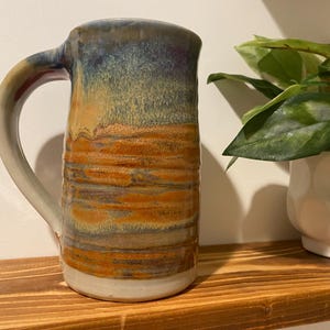 Può includere: Una tazza in ceramica fatta a mano con un grande manico. La tazza presenta una sfumatura di colore, che va dal blu in alto all'arancione e al marrone al centro, con una base bianca. La tazza è esposta su una mensola di legno.