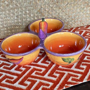 May include: A ceramic three-section serving dish featuring a vibrant orange and yellow gradient interior. The exterior is decorated with painted green leaves and purple accents, finished with a central purple handle shaped like a small vegetable.