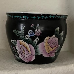 Peut inclure: Un pot de fleurs en céramique noire orné d'un motif floral peint à la main avec des roses roses et jaunes, des feuilles vertes et de petites fleurs bleues et violettes. Le bord présente un fin motif de vigne sarcelle sur fond noir.