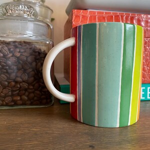 Puede incluir: Una taza de cer&aacute;mica con un asa blanca y un dise&ntilde;o a rayas de colores. Las rayas incluyen rojo, verde, azul y amarillo. La taza est&aacute; sobre una superficie de madera, con un tarro de granos de caf&eacute; en el fondo.