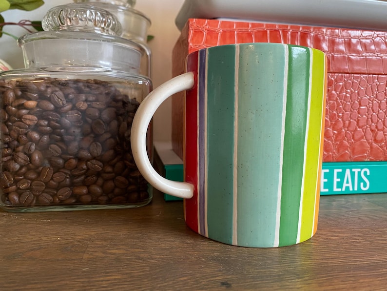 Puede incluir: Una taza de cer&aacute;mica con un asa blanca y un dise&ntilde;o a rayas de colores. Las rayas incluyen rojo, verde, azul y amarillo. La taza est&aacute; sobre una superficie de madera, con un tarro de granos de caf&eacute; en el fondo.