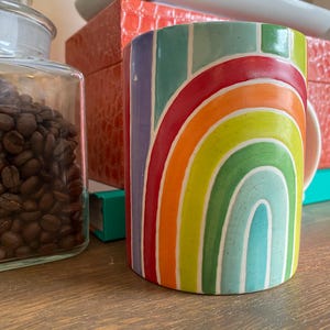 Puede incluir: Una taza de cer&aacute;mica cil&iacute;ndrica con un dise&ntilde;o de arco&iacute;ris. La taza presenta una vibrante gama de colores, incluyendo rojo, naranja, amarillo, verde y azul. La taza mide aproximadamente 10 cm de alto y est&aacute; sobre una superficie de madera.