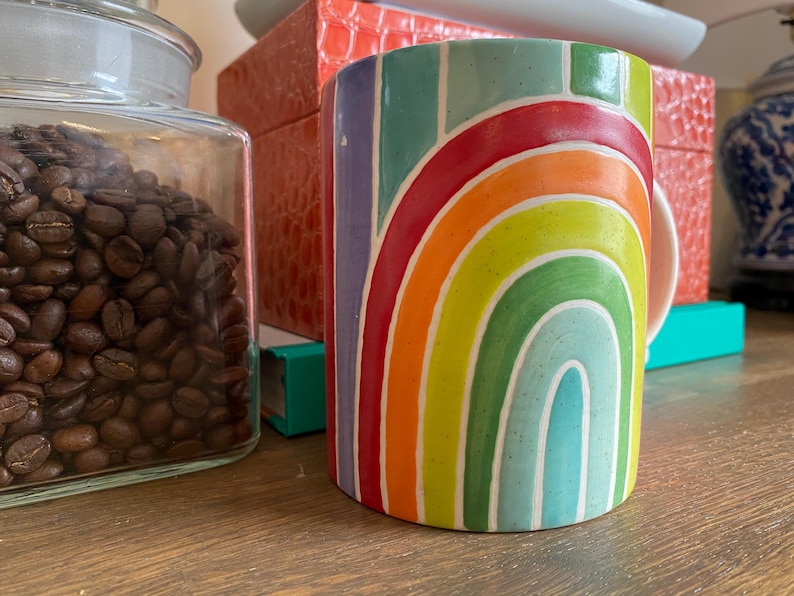 Puede incluir: Una taza de cer&aacute;mica cil&iacute;ndrica con un dise&ntilde;o de arco&iacute;ris. La taza presenta una vibrante gama de colores, incluyendo rojo, naranja, amarillo, verde y azul. La taza mide aproximadamente 10 cm de alto y est&aacute; sobre una superficie de madera.