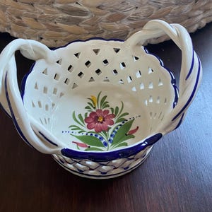 Op de afbeelding: Een witte keramische mand met een wand in rasterpatroon en twee gedraaide handvatten. De rand en handvatten zijn voorzien van blauwe accenten. De binnenkant toont een handgeschilderd bloemmotief met een roze bloem, groene bladeren en gele details.