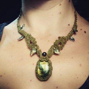 Può includere: Una collana in macramé verde con un grande ciondolo ovale in pietra verde e piccoli dettagli a forma di foglia.