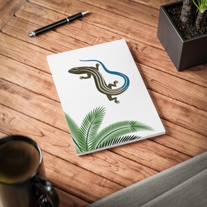 Puede incluir: Un cuaderno blanco con una ilustración de un lagarto azul y marrón y hojas de palmera verdes. El cuaderno está sobre una superficie de madera.