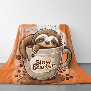 Op de afbeelding: Een oranje en bruine deken met een cartoon-luiaard in een koffiemok. De mok heeft de tekst "Slow Starter" erop.
